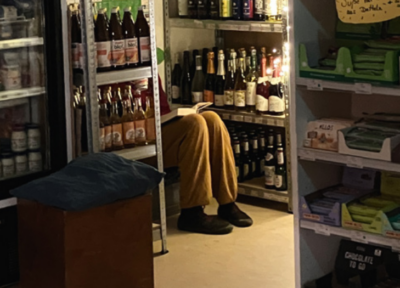 Das Bild zeigt jemanden der verstekct im Mila O in einer Ecke zwischen Getränkeregalen sitzt und liest