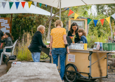 Das Bild zeigt 4 Personen die unter Wimpelketten stehen. Außerdem ist ein kleiner mobiler Essenswagen abgebildet.